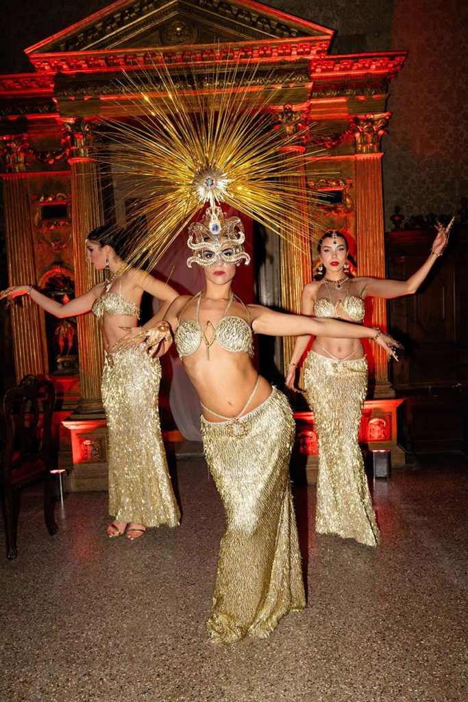 Three women in gold costumes and elaborate headdresses perform a dance before an ornate, gold-accented backdrop—an ideal moment for event videography to capture every dazzling detail.