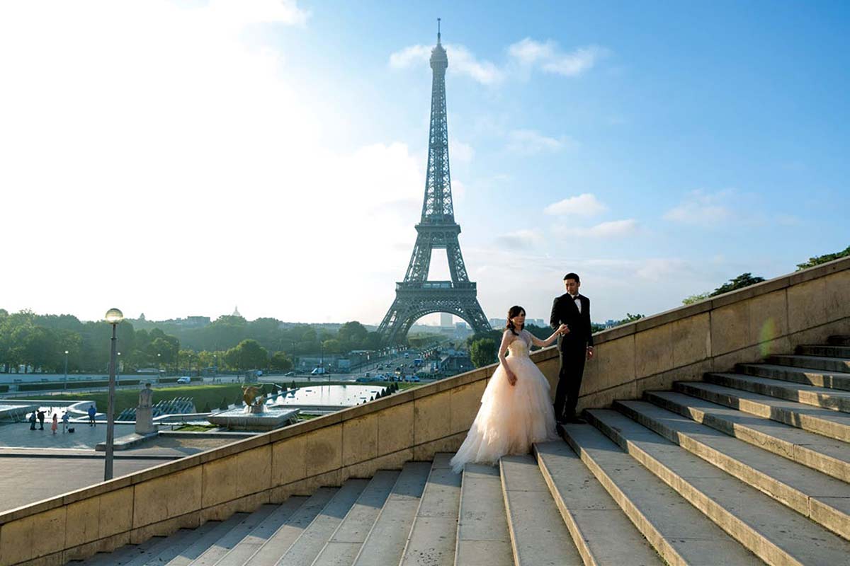 A couple in formal wedding attire stands on outdoor steps with the Eiffel Tower in the background on a clear day- filmed by Luxury Wedding Videographer Story Of Your Day