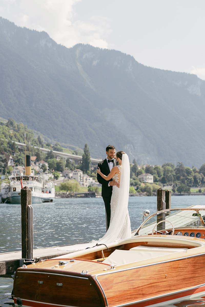 A bride and groom embrace on a wooden dock by a lake, with a boat in the foreground and mountains in the background