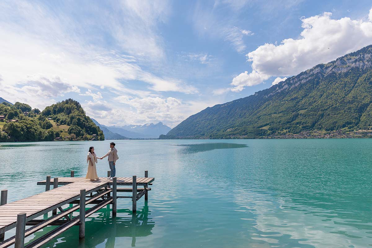 A couple stands on a wooden dock over a turquoise lake, surrounded by mountains and a cloudy blue sky
