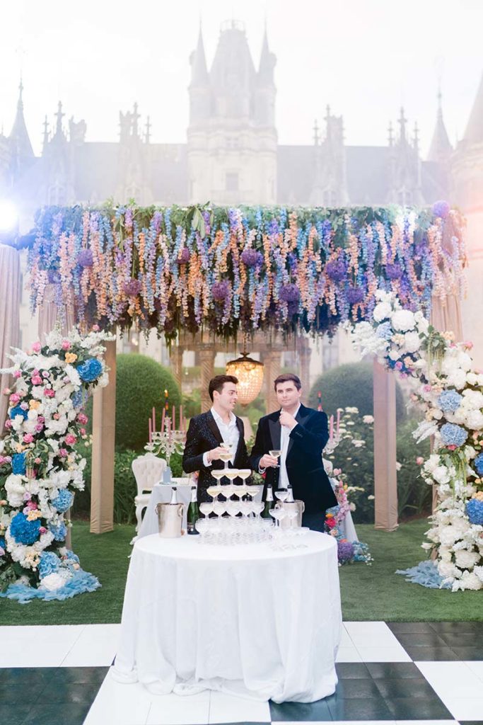 Two men stand by a champagne tower at an outdoor event with floral decorations and a grand building in the background, perfect for a draft home page showcasing elegant celebrations.