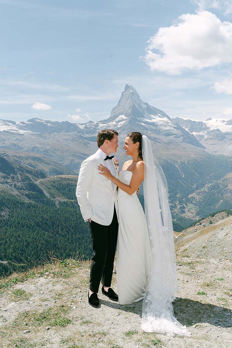 A bride and groom stand together outdoors in wedding attire, with mountains and the Matterhorn visible in the background under a blue sky, perfect for a draft home page capturing a romantic alpine celebration.