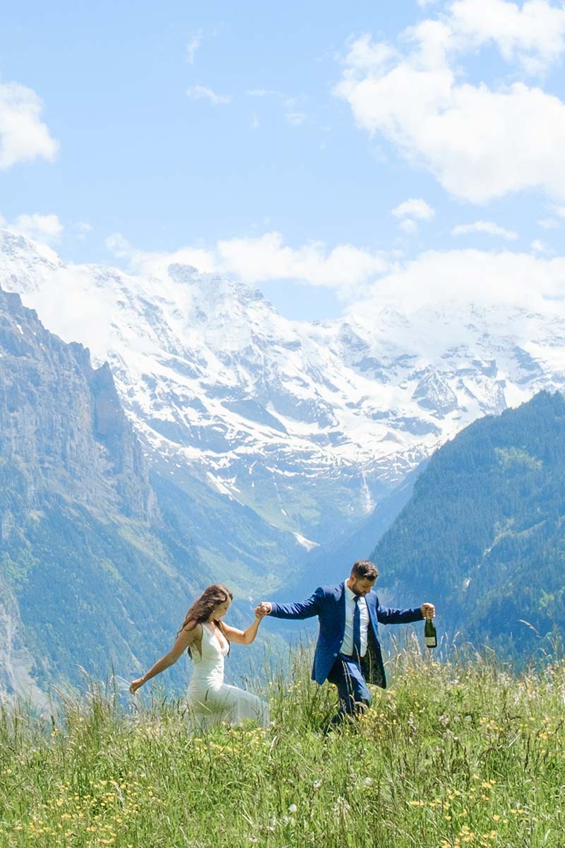 A couple dressed in formal attire walks through a grassy field with mountains and snow-capped peaks in the background, capturing the elegance perfect for a draft home page. The man holds a bottle in one hand.
