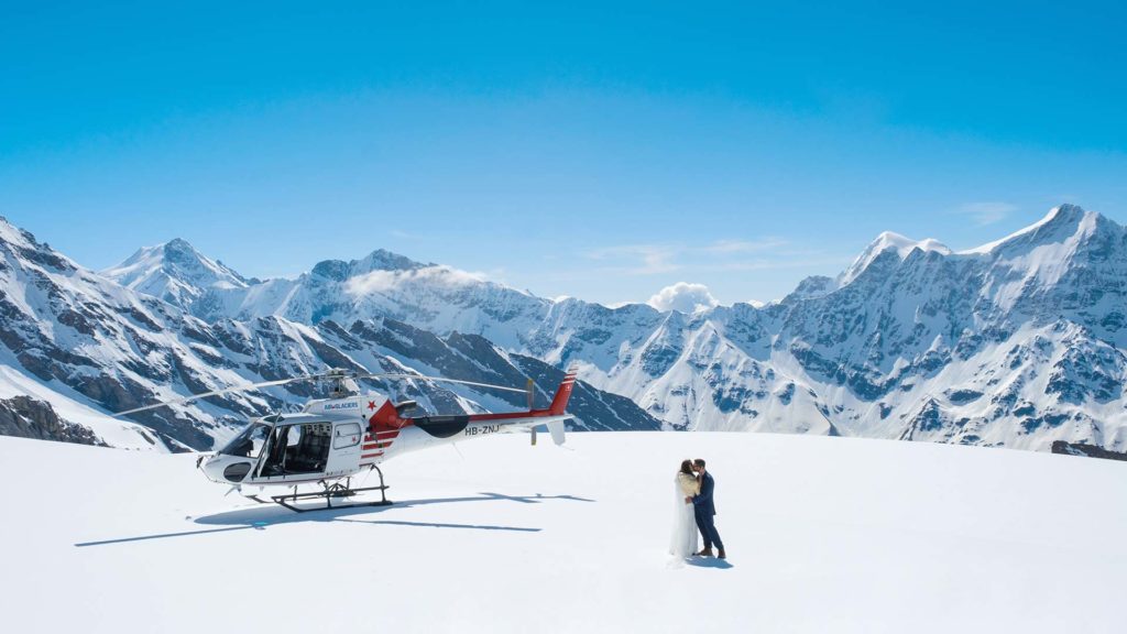 A couple in wedding attire stands on a snowy mountain next to a helicopter, beneath a clear blue sky—perfect inspiration for your draft home page with breathtaking, adventure-filled romance.