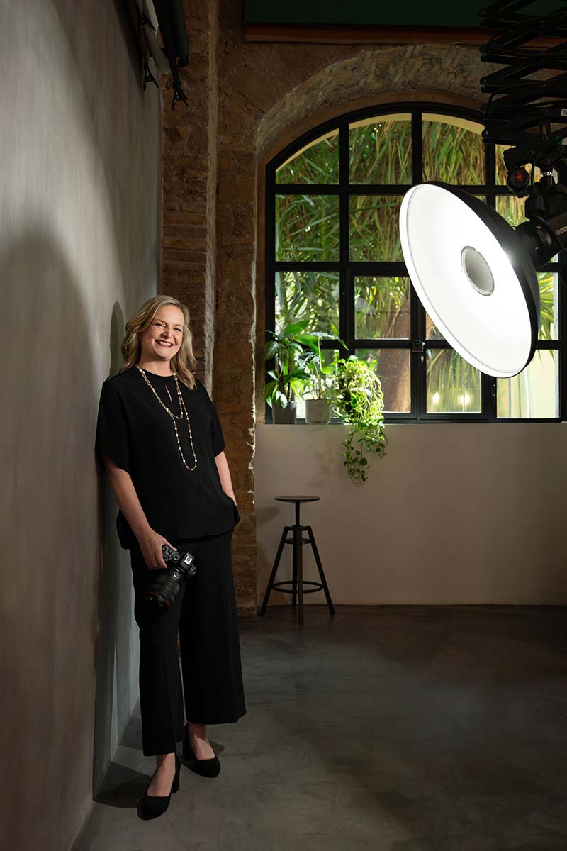 A woman in black clothing stands against a wall in a photography studio, holding a camera. The DRAFT HOME PAGE shoot features a large softbox light and a window with plants in the background.