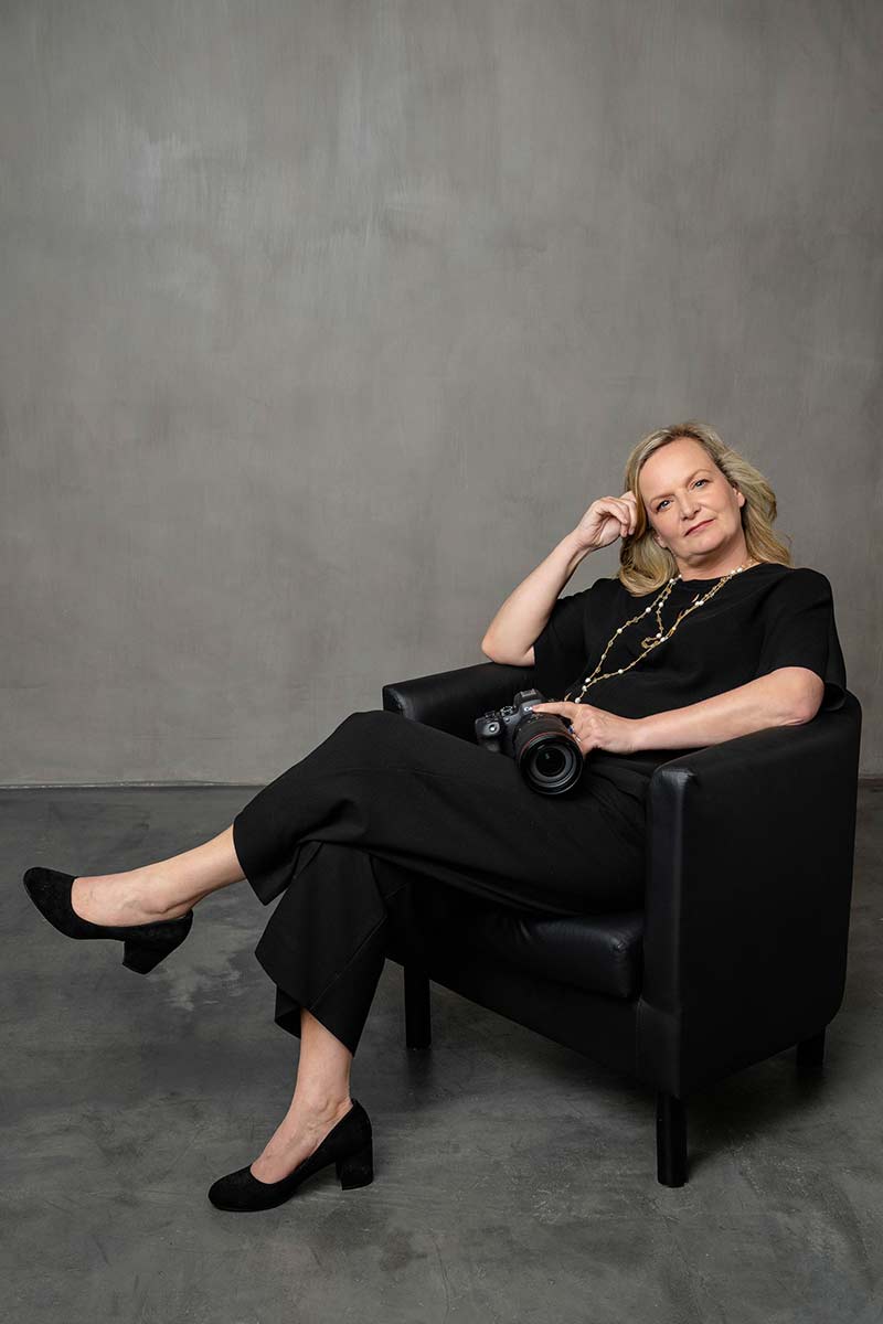 A woman dressed in black sits in a black armchair, holding a camera, ready to capture unforgettable weddings, against a plain gray background.