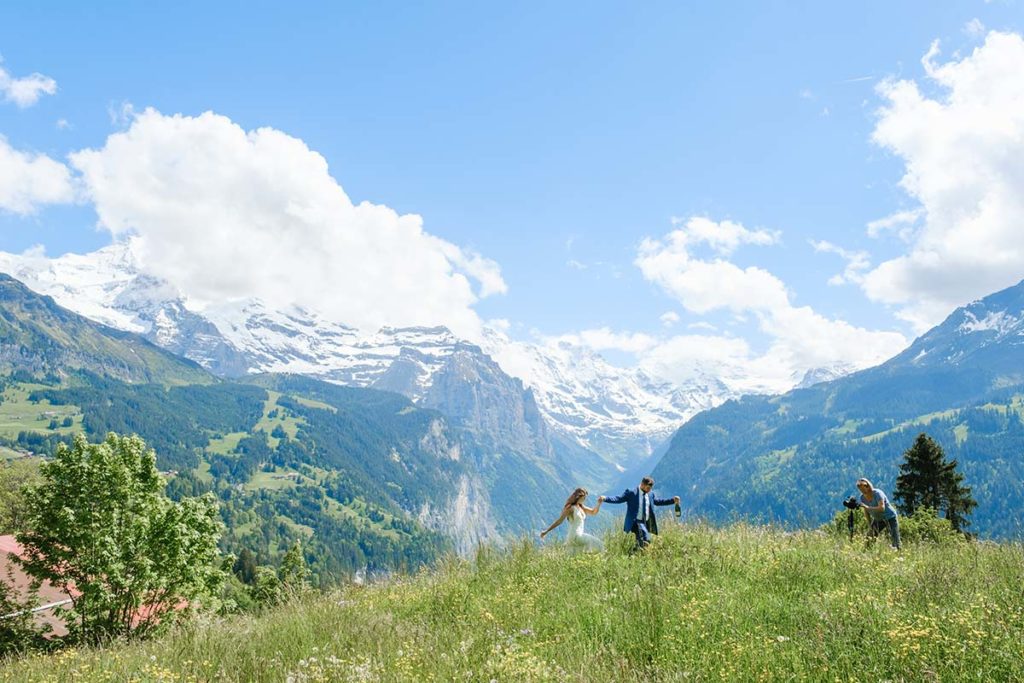 Four people walk through a grassy meadow, perhaps heading to a weddings celebration, with snow-capped mountains and a blue sky with clouds in the background.