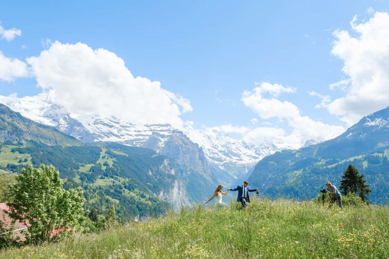 Four people walk through a grassy meadow, perhaps heading to a weddings celebration, with snow-capped mountains and a blue sky with clouds in the background.