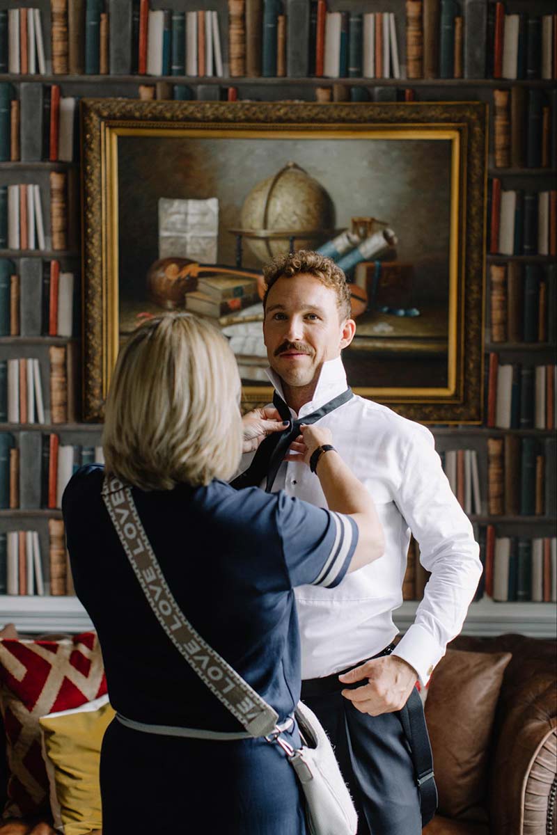A woman helps a man adjust his black bow tie in a room with book-patterned wallpaper and a framed painting—capturing a quiet moment of preparation before weddings.