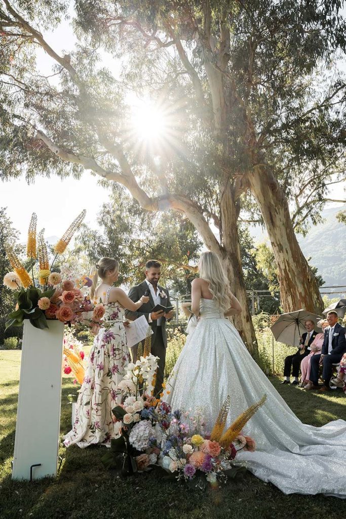 A bride and groom stand with an officiant at an outdoor weddings ceremony, surrounded by flowers and trees, with sunlight streaming through the branches.