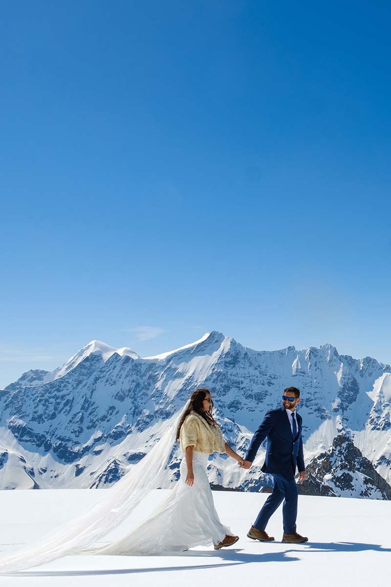 A bride and groom walk hand-in-hand on snow with a backdrop of snow-covered mountains under a clear blue sky, capturing the magic of winter weddings.