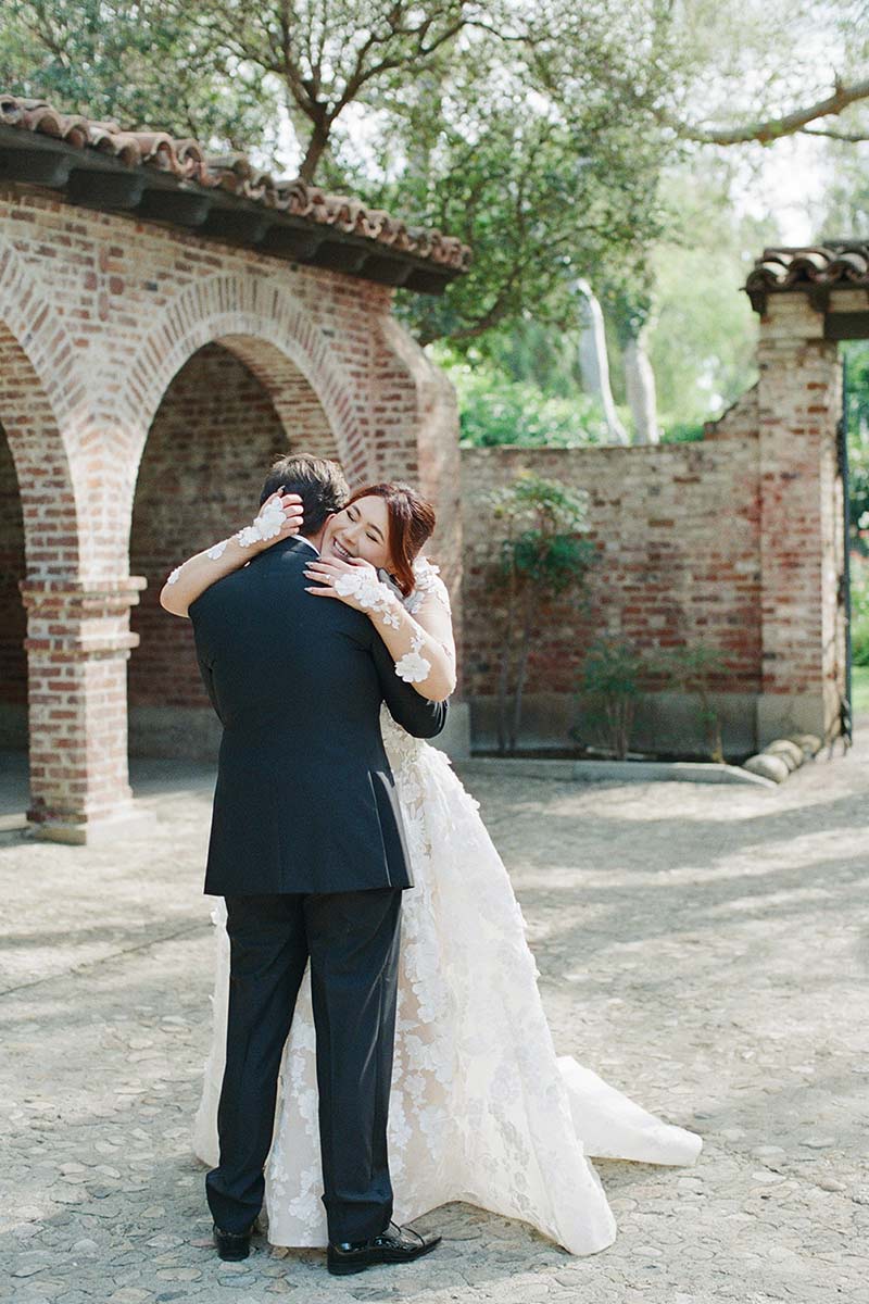 The bride and groom share an embrace outdoors beside a brick building with arches, surrounded by trees and sunlight, capturing the magic of weddings in a perfect moment.