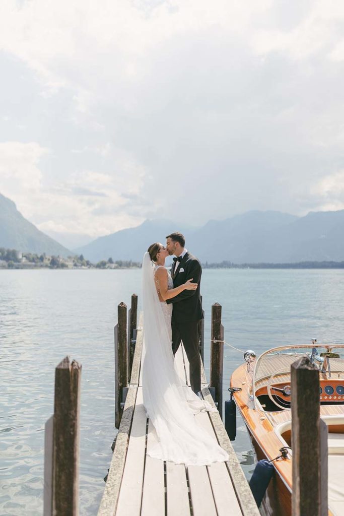 A bride and groom share a kiss on a wooden dock beside a boat, with mountains and a lake in the background—a perfect moment from their unforgettable weddings day.