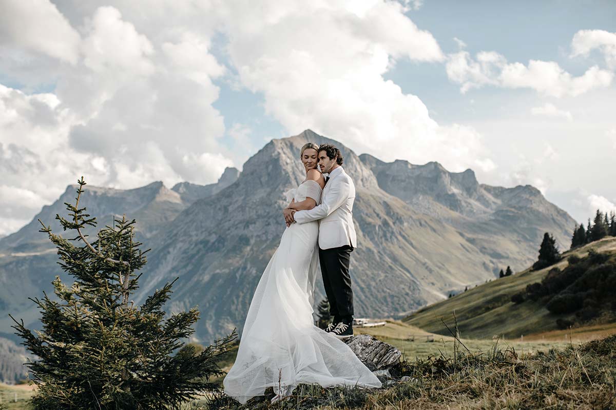 A bride and groom embrace outdoors on a grassy hillside with mountains and clouds in the background, evoking the timeless romance often seen in Austrian cinema.