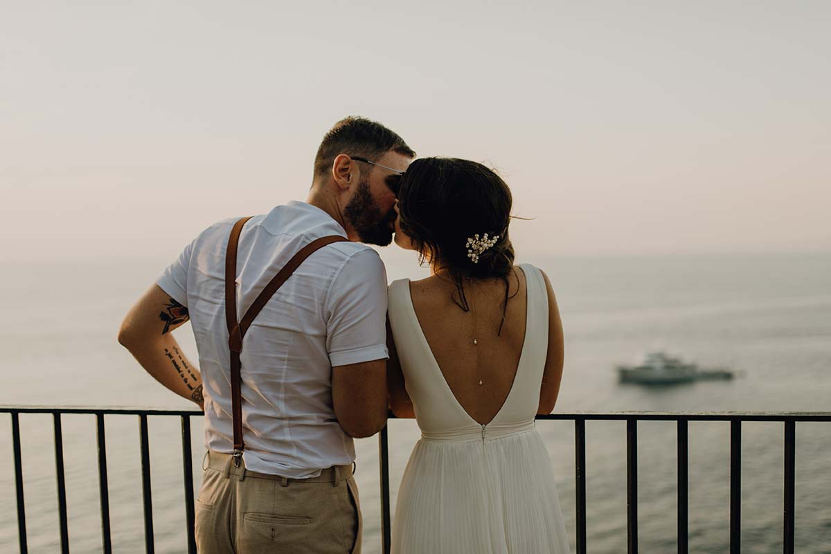 A couple dressed in wedding attire stands on a balcony overlooking the ocean at sunset, with a boat visible in the water below—capturing a scene reminiscent of classic Italian Cinema.