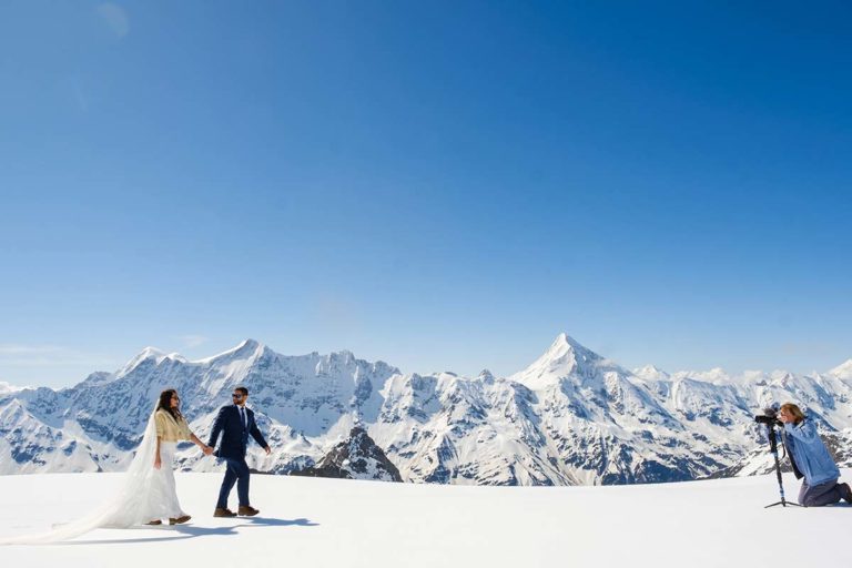 A bride and groom walk hand in hand on snowy ground with a photographer capturing them; snow-covered mountains and a clear blue sky are in the background.