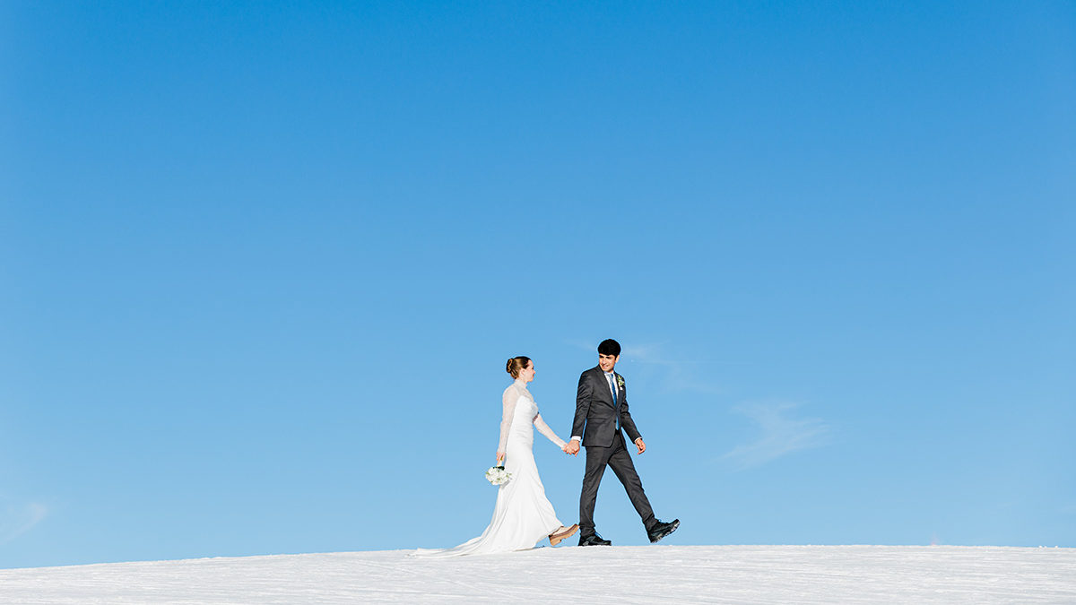 A bride and groom hold hands while walking on snow under a clear blue sky on the top of Männlichen above Wengen during their Swiss Mountain elopement.