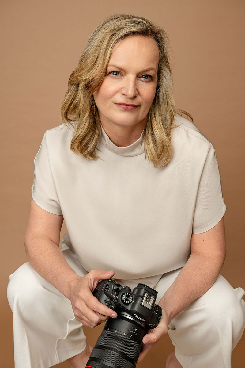 A woman with blonde hair in a light outfit sits holding a professional camera against a plain beige background, capturing the essence of wedding videography in France.