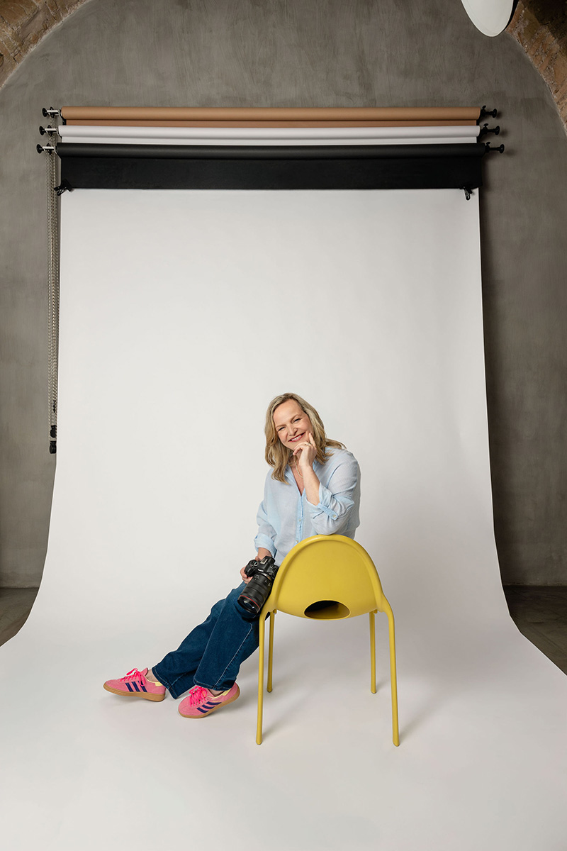 Emma Wilson sits on a backward yellow chair in front of a photography backdrop, holding a camera and smiling—ready to capture Italy’s charm through wedding videography. She wears jeans, a light blue shirt, and bright pink sneakers.