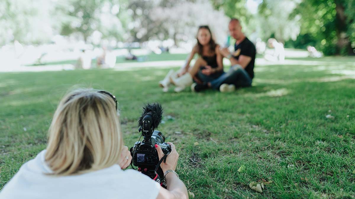 A person lies on the grass taking a photo or video of two people sitting together in a park on a sunny day.