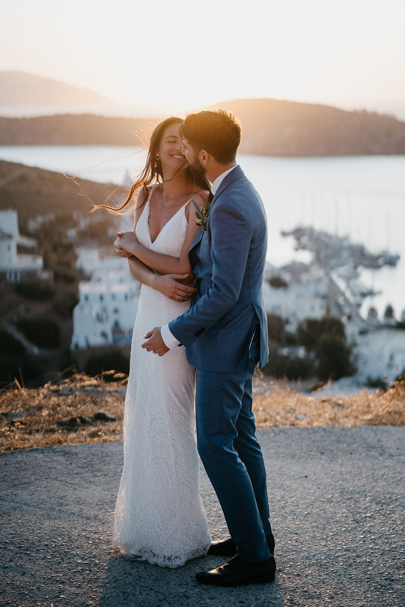 A bride in a white dress and a groom in a blue suit smile and embrace outdoors at sunset, with a coastal village and boats in the background—a perfect scene for beautiful destination wedding videography.