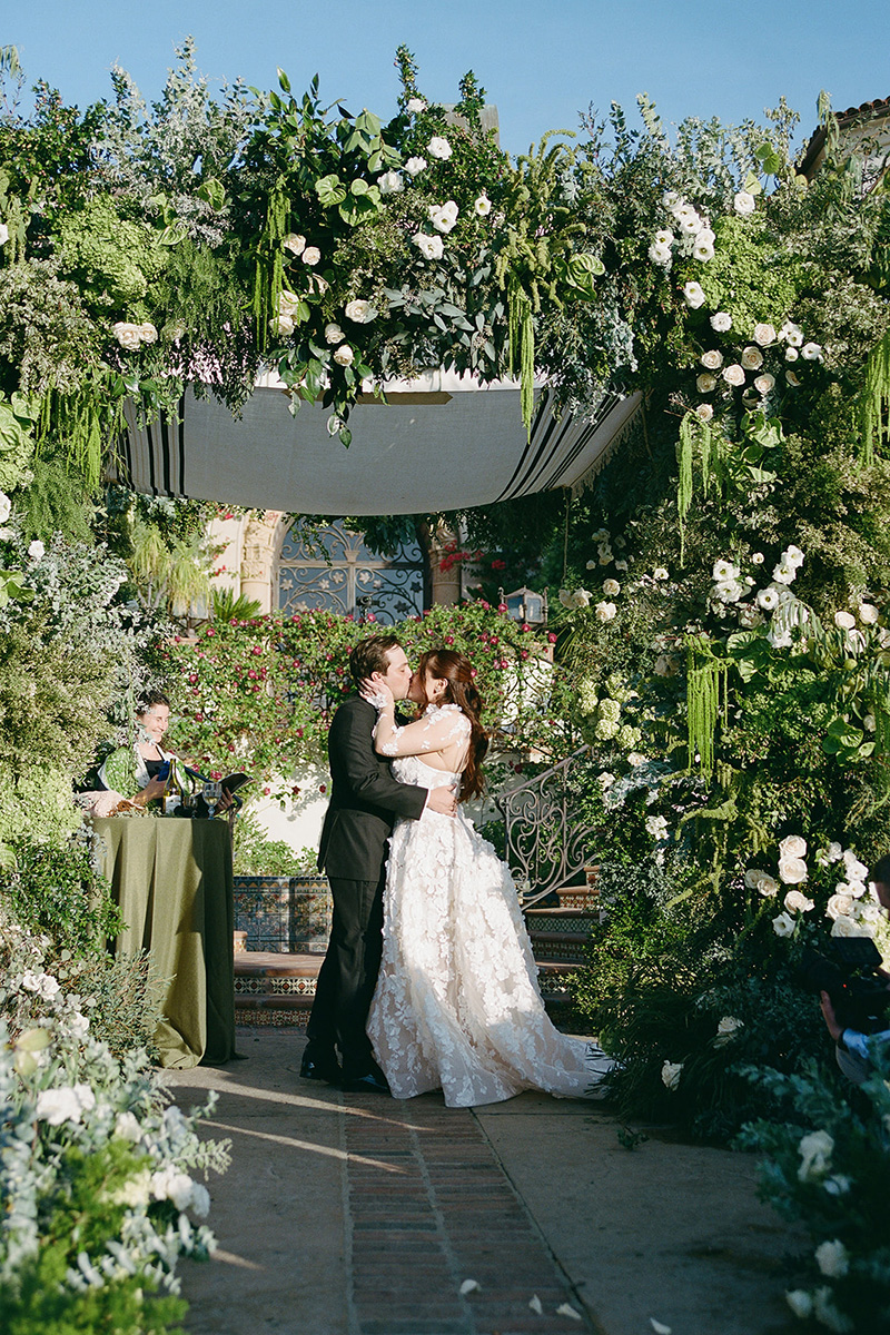 A bride and groom share a kiss under a lush, flower-filled arch at an outdoor destination wedding ceremony, with a person standing nearby at a table—perfect for capturing unforgettable wedding videography moments.