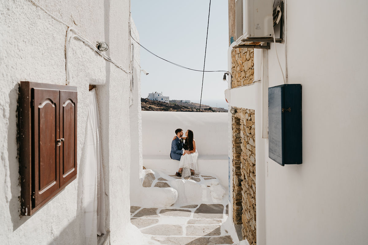 A couple sits close together on a white stone ledge in a narrow alley with whitewashed walls, under bright daylight in a Mediterranean setting—an idyllic scene perfect for destination wedding videography.