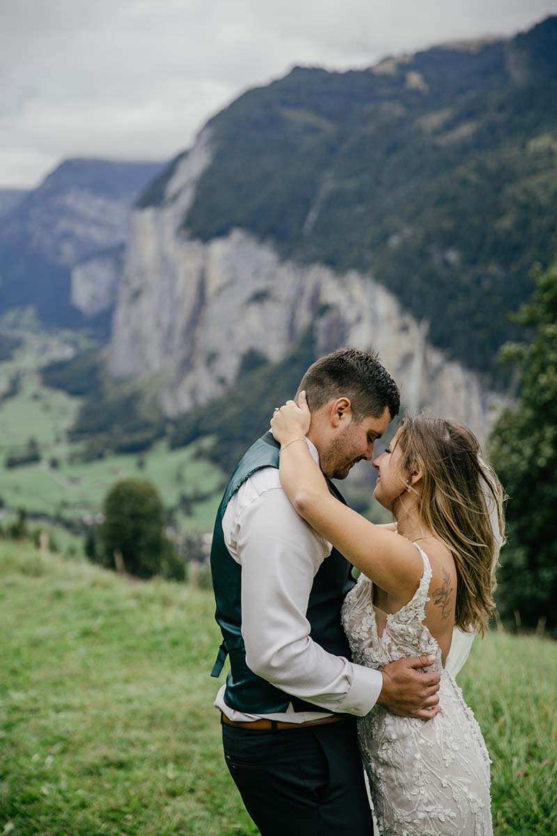 A couple in wedding attire embraces on a grassy hillside with mountains and cliffs in the background under a cloudy sky, captured beautifully Emma Wilson from Story Of Your Day
