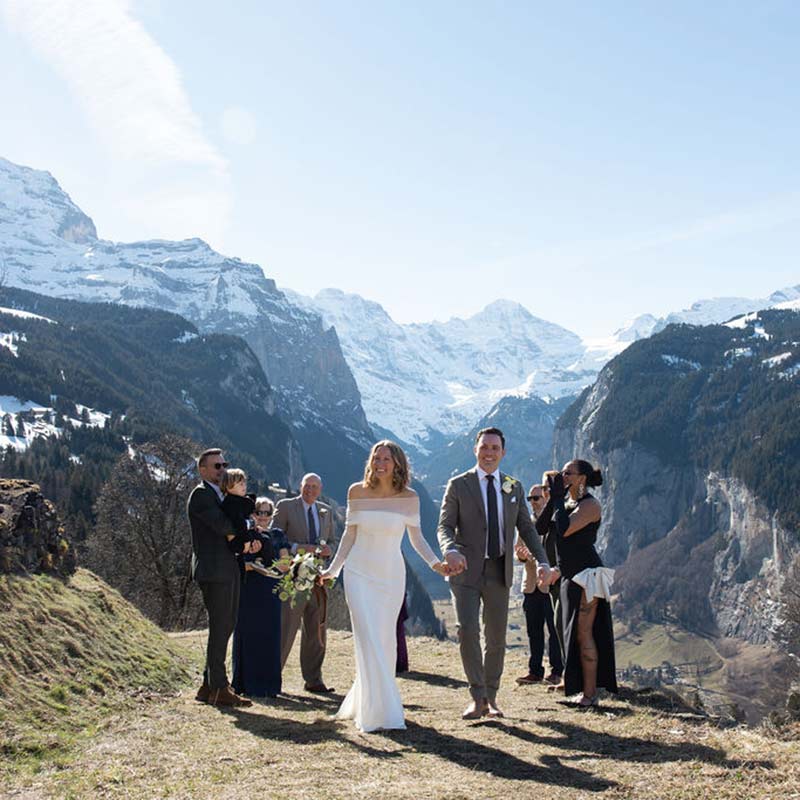 A bride and groom walk hand in hand outdoors, surrounded by a small group of people and stunning snow-capped mountains—perfect for a small wedding videographer to capture every intimate moment.