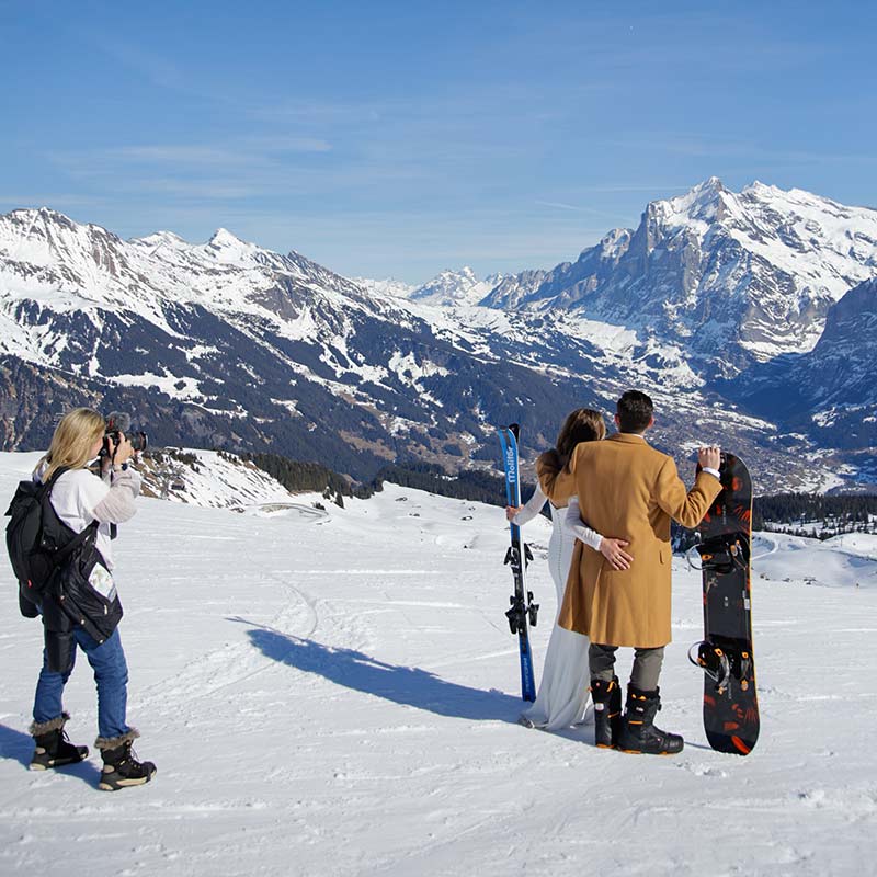 Emma Wilson from Story Of Your Day films a couple in wedding outfits holding skis and a snowboard, standing on a snowy mountain with a scenic alpine backdrop