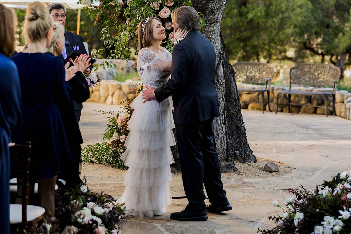 A bride and groom stand close together outdoors during their wedding ceremony in Santa Barbara surrounded by guests and floral decorations.