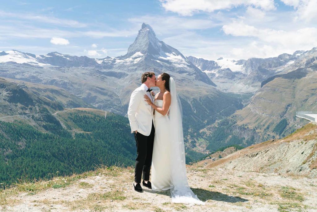 A bride and groom kiss outdoors on a mountain overlook, with the Matterhorn visible in the background under a partly cloudy sky—a perfect moment for Wedding Videography Switzerland.