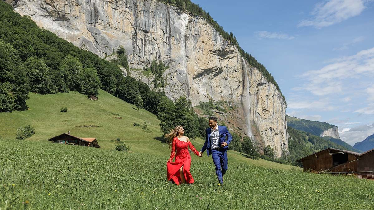 A woman in a red dress and a man in a blue suit walk hand in hand across a grassy field with cabins and a rocky cliff behind them—a perfect moment for any proposal photography or photography portfolio.