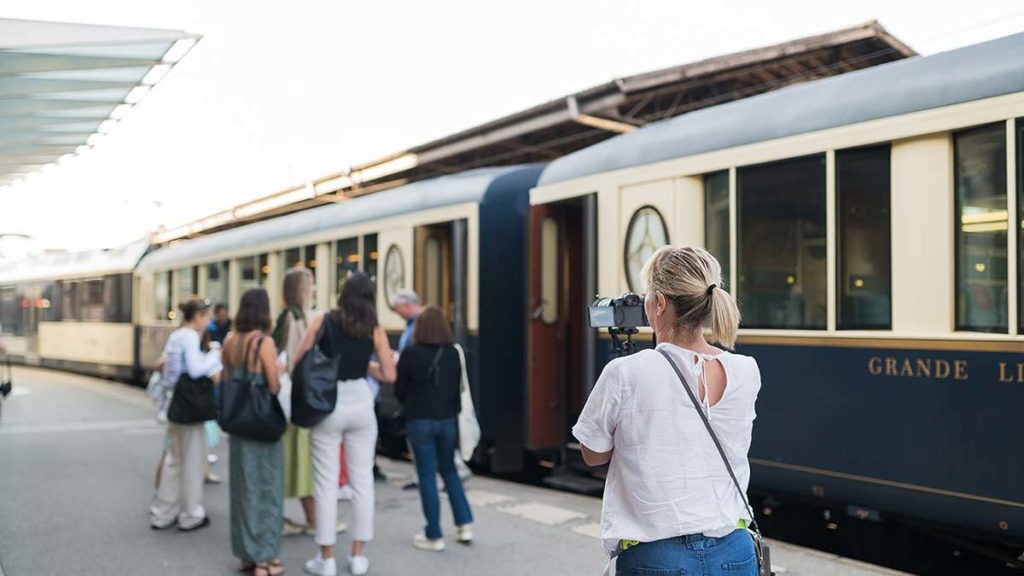A group of people stand on a train platform near a blue and cream-colored train, while one person takes a photo of them.