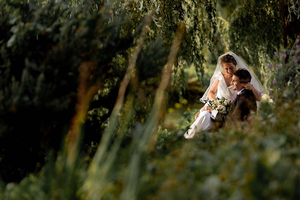 A bride and groom sit closely together outdoors, surrounded by greenery and soft sunlight, with the bride holding a bouquet.