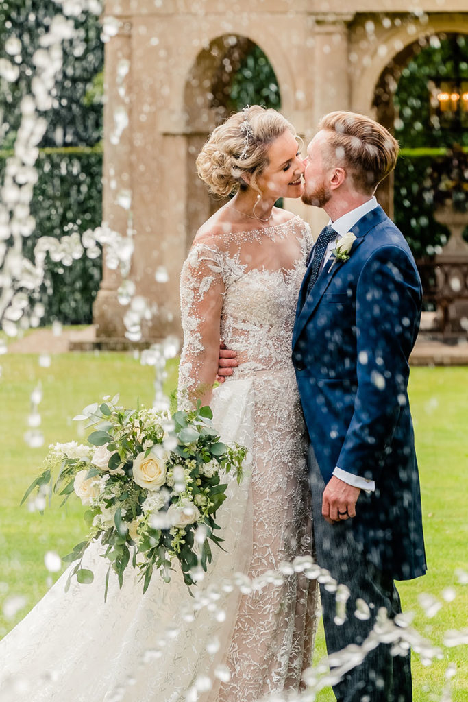 A bride and groom kiss outdoors before a stone building, viewed through streams of water from a nearby fountain. The magical moment is perfect for stunning wedding videography by a talented UK wedding videographer.