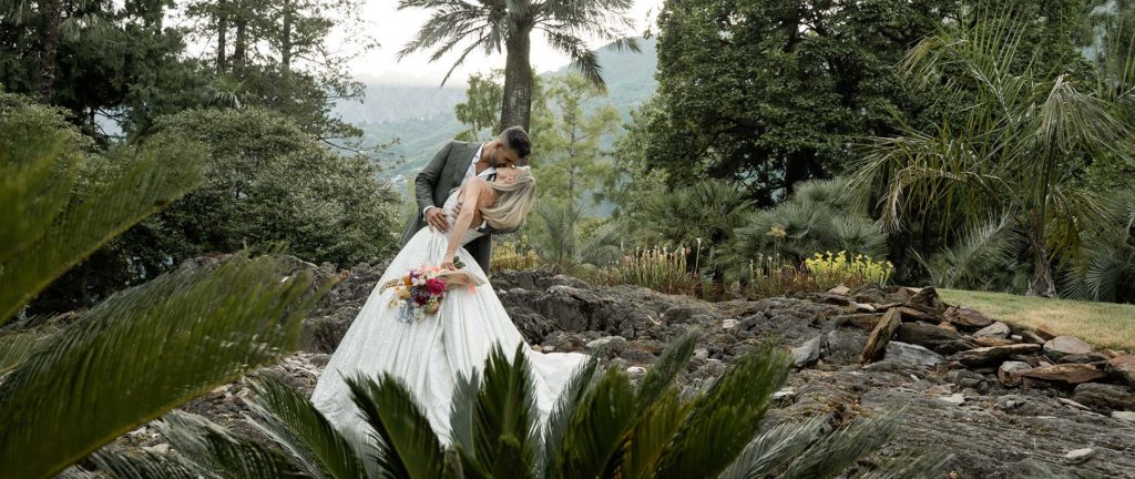 A bride and groom kiss outdoors on rocky ground, surrounded by lush green trees and plants, with mountains visible in the background.