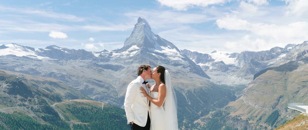 A bride and groom kiss outdoors with snow-capped mountains and the Matterhorn in the background under a partly cloudy sky.