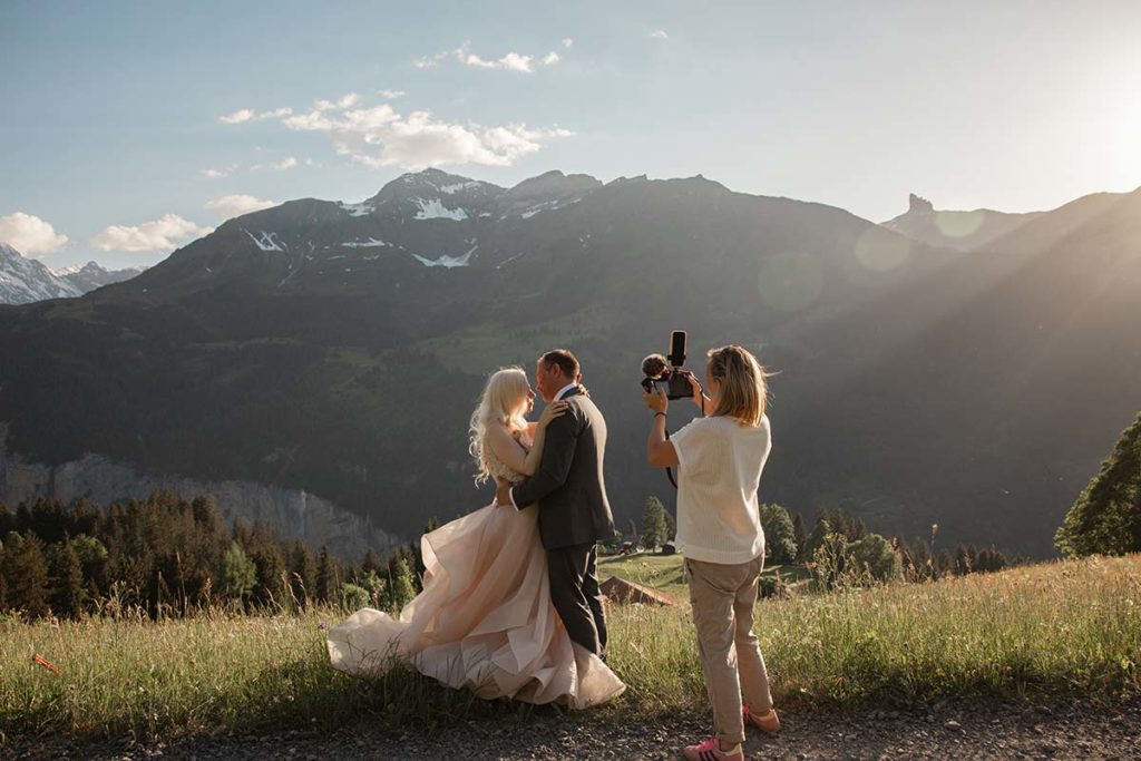 A couple in wedding attire poses for photographer Emma Wilson on a grassy hillside in Wengen with mountains and sunlight in the background