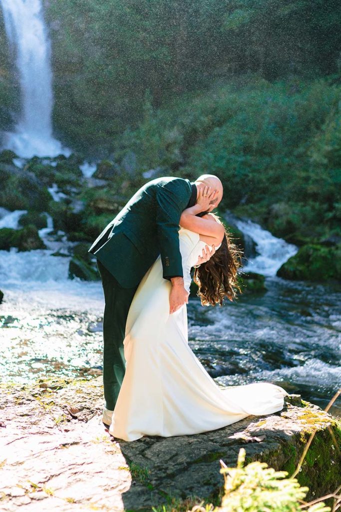 A couple in wedding attire kisses on a rocky surface near a waterfall, surrounded by greenery and flowing water—capturing the essence of adventurous elopement planning.