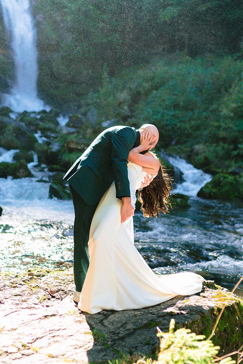 A couple in wedding attire kisses on a rocky surface near a waterfall, surrounded by greenery and flowing water capturing the essence of adventurous elopement planning.
