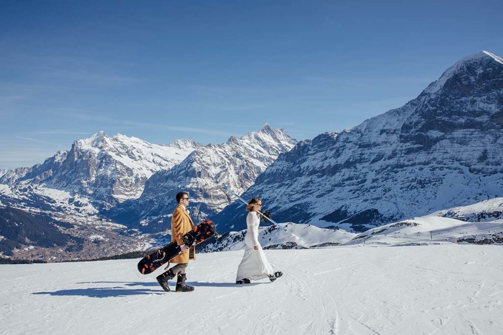 Two people, one in a brown coat and the other in a white wedding dress, walk on a snowy mountain slope carrying snowboards an adventurous elopement set against snow-covered peaks and a clear sky.