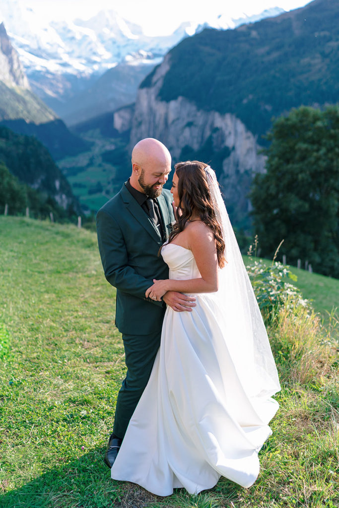 A bride and groom stand close together outdoors on a grassy Wengen hilltop, with mountains and a valley in the background capturing the perfect moment for an unforgettable elopement.