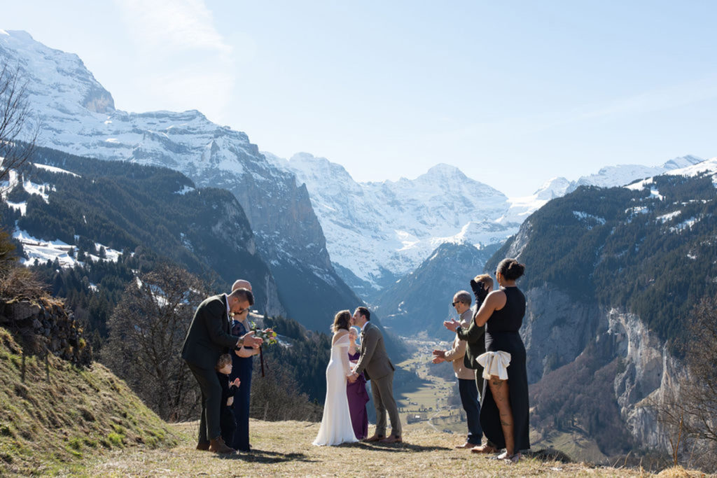 Three couples stand on a grassy hillside with snow-capped mountains in the background; two couples kiss while others watch and clap, capturing a joyful elopement moment perfect for your Elopement Page.