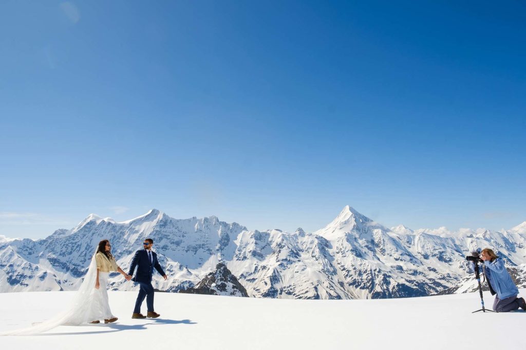 A couple dressed in wedding attire holds hands on a snowy mountain plateau, embracing their elopement as a photographer captures the moment; snow-covered peaks are visible in the background under a clear blue sky.