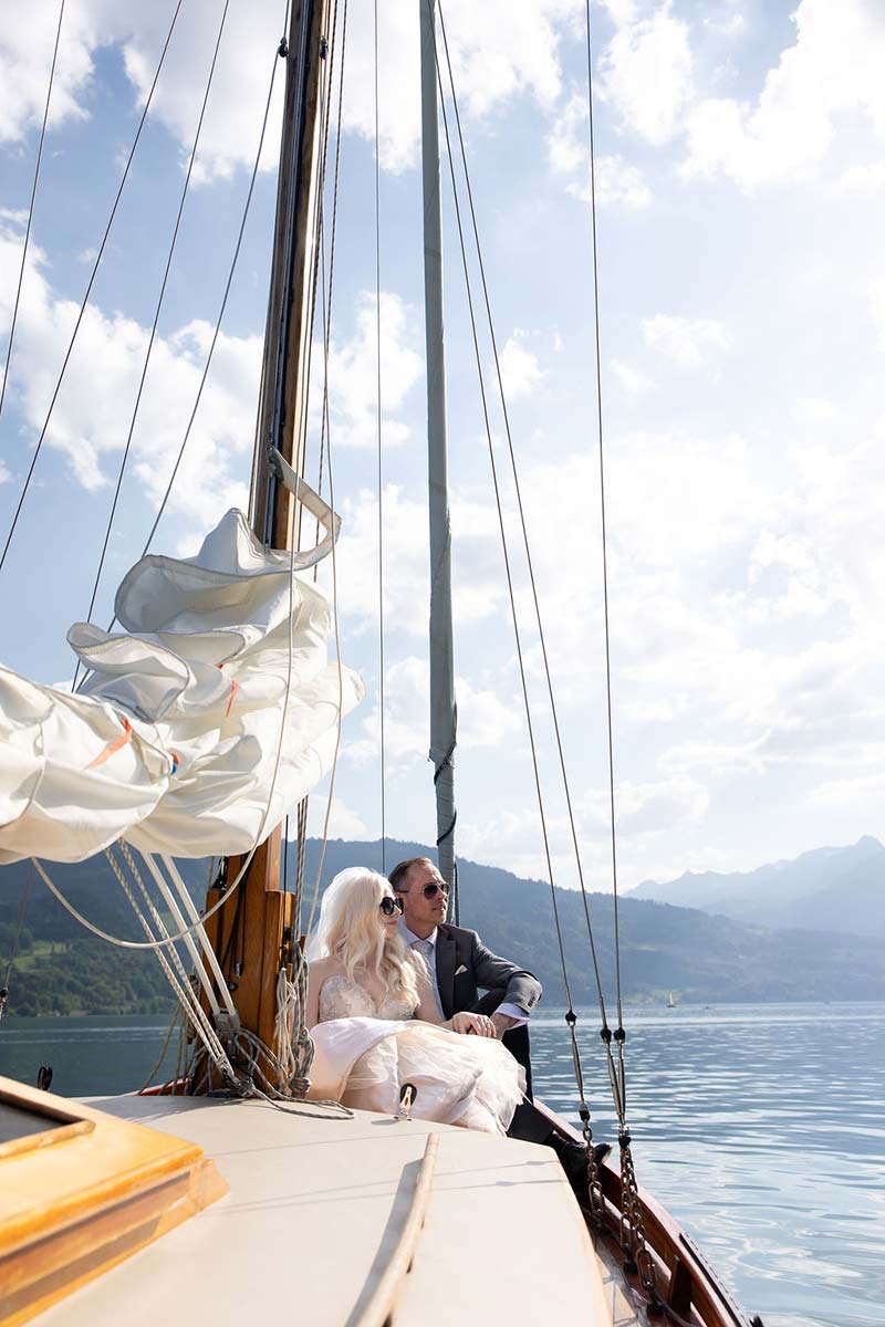 A couple in formal attire sits together on the deck of a sailboat on a calm lake, with mountains and a partly cloudy sky—an intimate elopement captured against nature’s breathtaking backdrop.