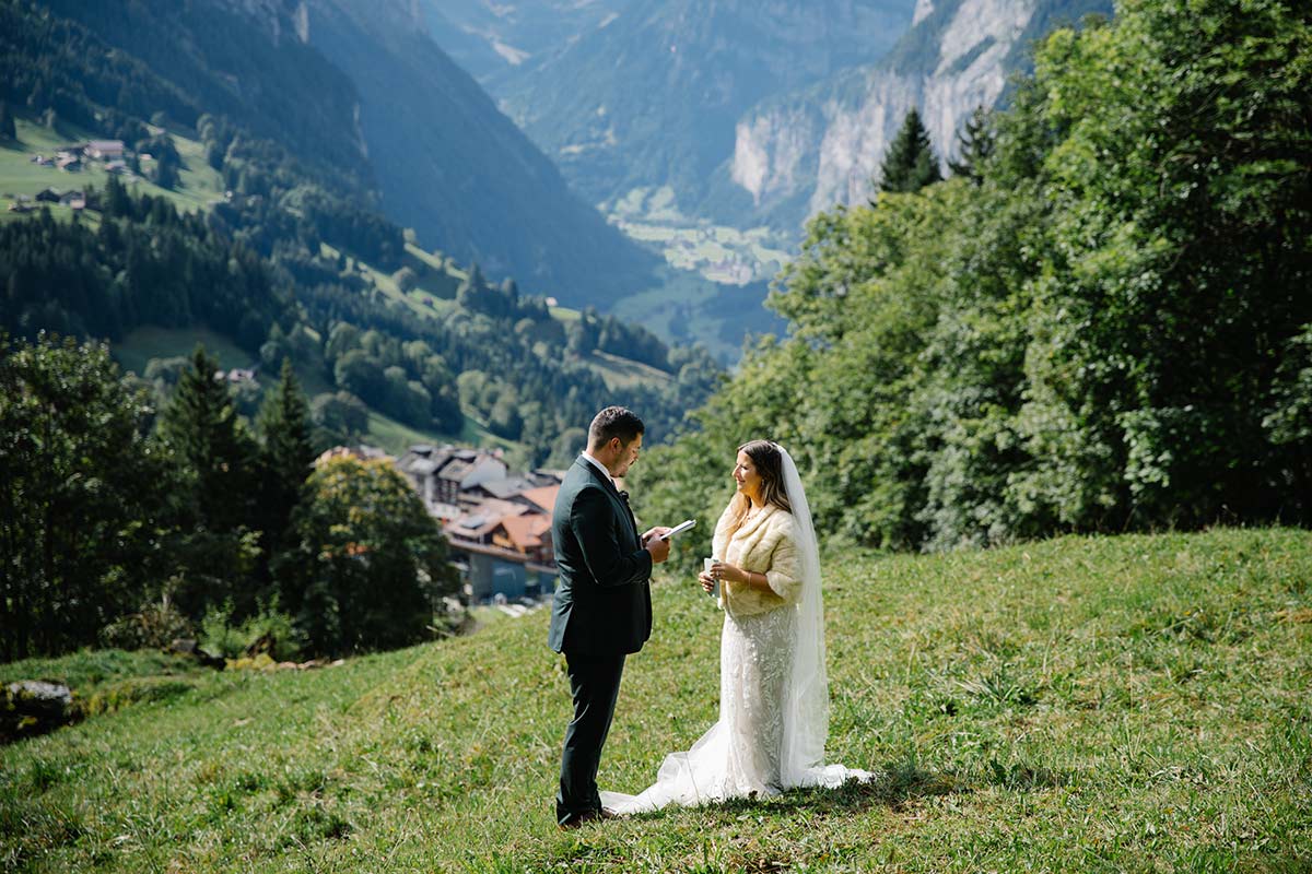 A bride and groom stand on a grassy hillside, exchanging vows with a village and mountains in the background—an unforgettable moment perfect for any Elopement Page.