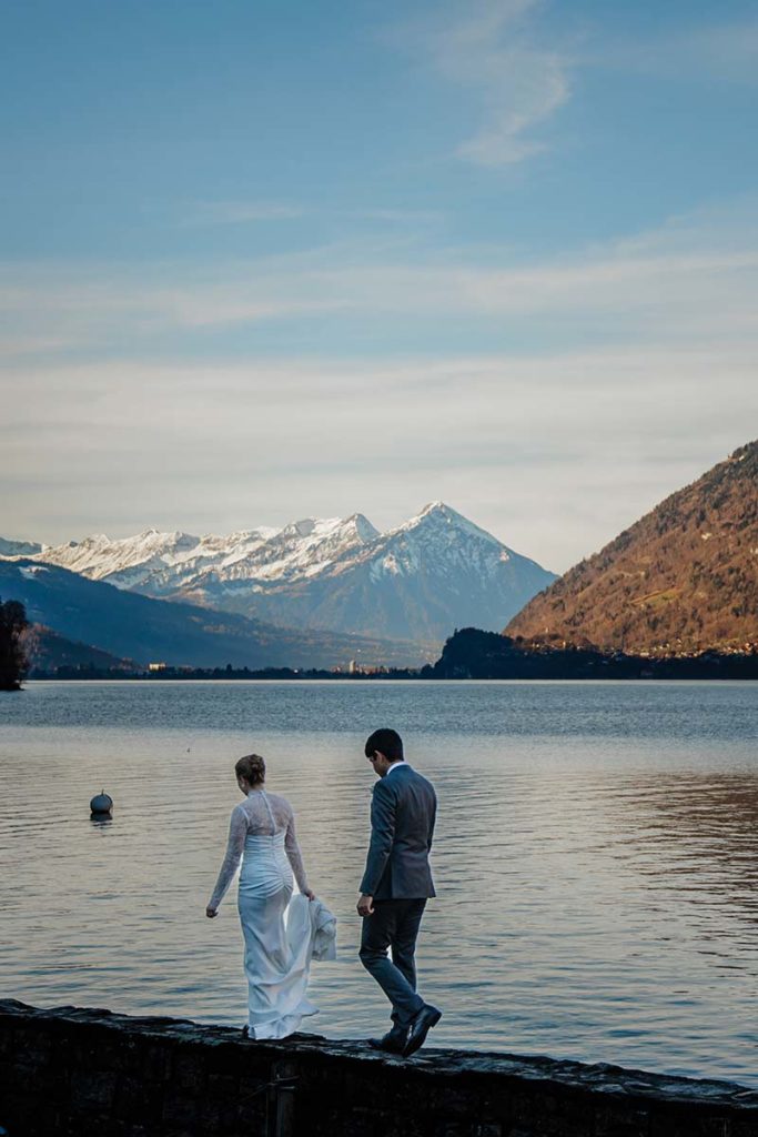 A couple in wedding attire walks by a lake with snow-capped mountains and a clear sky—a perfect elopement scene.