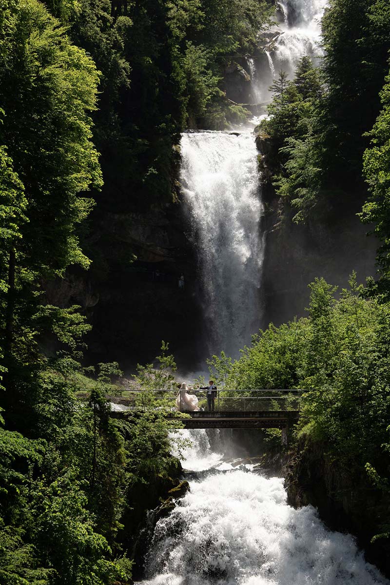 The Geissbach falls flows through dense green forest, with an elopement couple standing on a wooden bridge