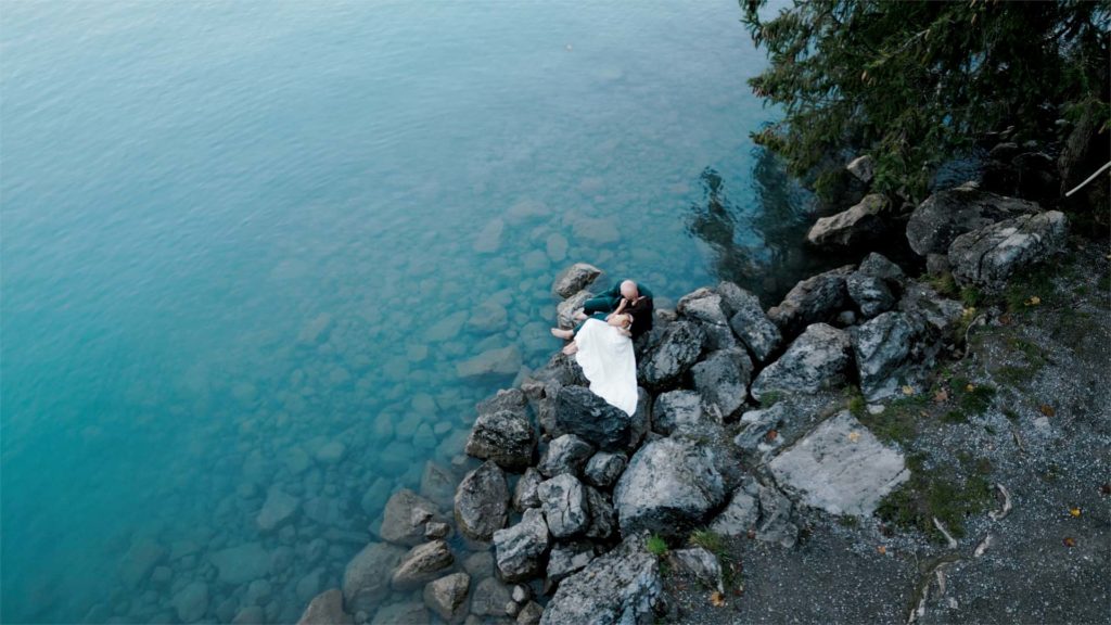 A couple celebrates their elopement in formal attire, lying on large rocks beside clear blue water, surrounded by trees and shoreline.